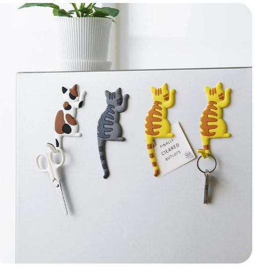 Set of four cat-shaped hooks on a white surface with a plant in the background.