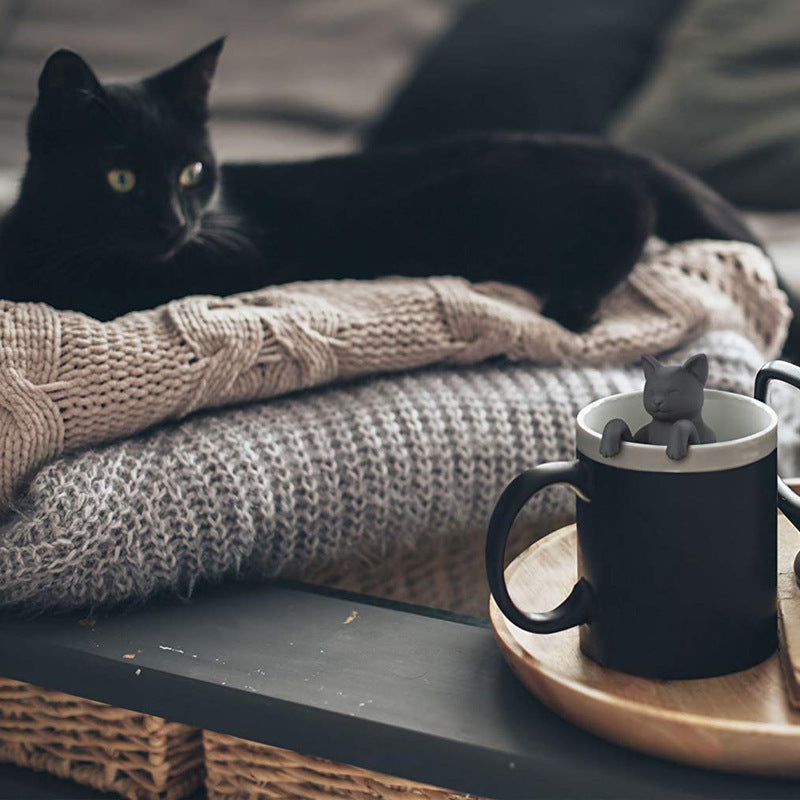 Black cat lying on a blanket with a mug featuring a small cat figure on a wooden tray.