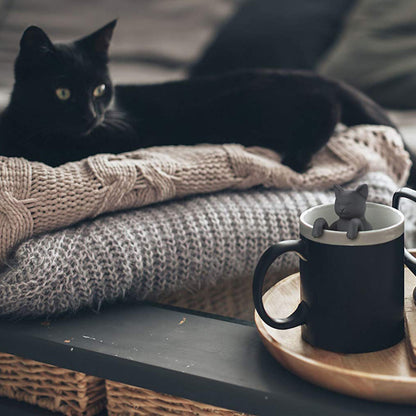 Black cat lying on a blanket with a mug featuring a small cat figure on a wooden tray.