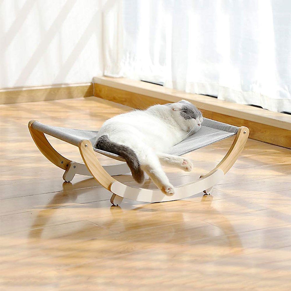 Cat resting on a small, elevated wooden platform with a soft cushion in a room with light-colored curtains.