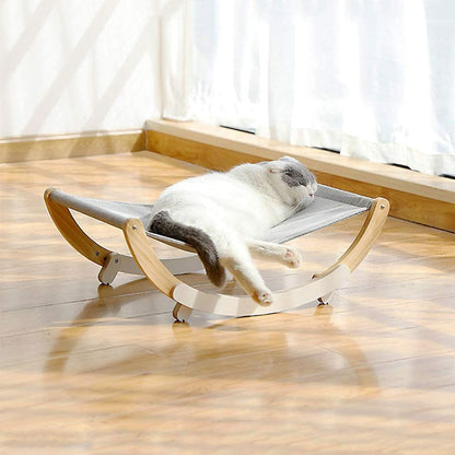 Cat resting on a small, elevated wooden platform with a soft cushion in a room with light-colored curtains.